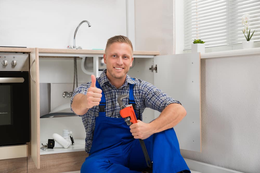 Happy plumber with an adjustable wrench sitting in a kitchen